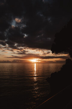 Dramatic sunset over the ocean with dark storm clouds, golden sunlight reflecting across calm water, and a peaceful coastal horizon at dusk.