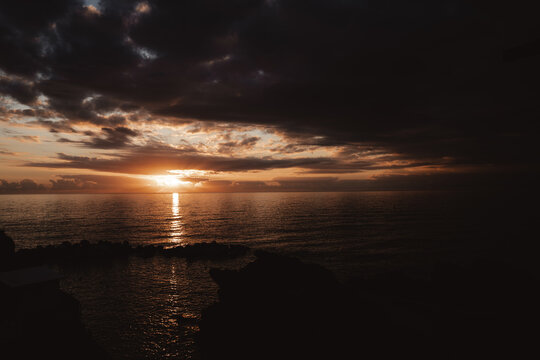Dramatic sunset over the ocean with dark storm clouds, golden sunlight reflecting across calm water, and a peaceful coastal horizon at dusk.