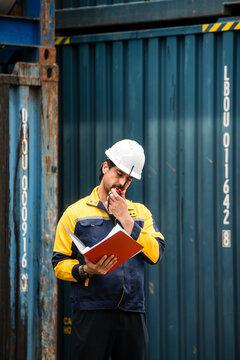 A hard-hat worker reviews documents beside stacked shipping containers, planning logistics and safety checks at a freight yard.
