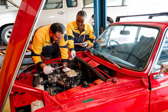 Two auto mechanics in yellow uniforms service the engine of a classic red car inside a repair shop.