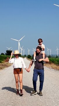 Family walks a gravel road past wind turbines on a sunny day; child rides on shoulders as blue sky and green fields frame outdoor leisure and clean energy
