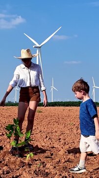 Family plants a young tree in a dry field under blue sky, with wind turbines in the background, highlighting sustainability and renewable energy.