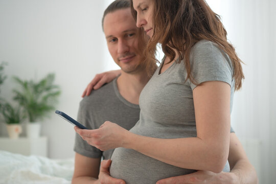 Pregnant woman and husband standing together in bright bedroom sharing pregnancy news on a phone call. Expecting mother with arm around waist while partner holds phone communicating with family.