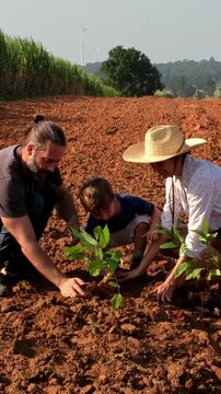 Two adults and a child plant seedlings in a red-soil field under a sunny sky, with wind turbines on the horizon-teamwork for sustainable farming.