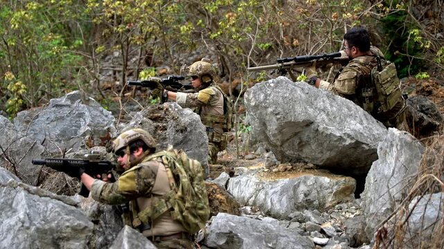 Three armed soldiers in camouflage take cover among large rocks, aiming rifles during a tactical operation in rugged, forested terrain.