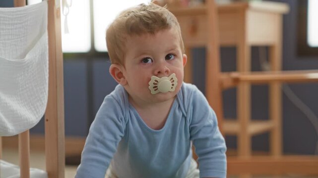 Toddler boy with pacifier crawling on nursery floor, face up and gaze toward window by wooden crib in home building; curiosity exploration comfort.