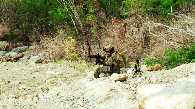 A camouflaged soldier kneels in a rocky riverbed, aiming a rifle toward dense scrub, using the terrain for cover during a tactical patrol or training exercise.