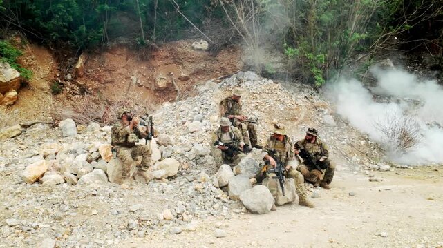 Camouflaged soldiers take cover on a rocky hillside during a field exercise, rifles ready as smoke drifts across the terrain.