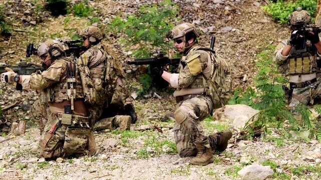 Armed soldiers in camouflage move through rocky terrain, aiming rifles and coordinating as a tactical team during a field training exercise.