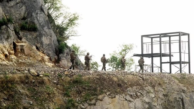 A group of soldiers in camouflage patrol along a rocky cliffside near a metal scaffolding structure, with sparse trees clinging to the rugged slope under an overcast sky.