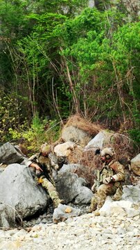 Two camouflaged soldiers rest among large rocks at the edge of a dense forest, communicating during a tactical stop in rugged terrain.