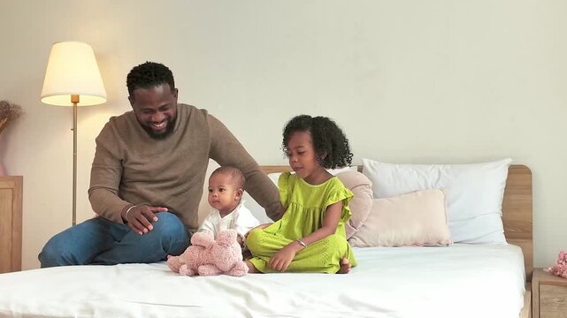 Father sits on a bed with his daughter and baby, playing with a pink teddy bear in a bright, cozy bedroom