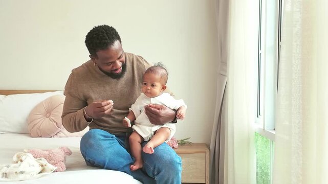 A loving parent sits on a bed by a bright window, gently holding a baby in a cozy bedroom, sharing a quiet moment of bonding and care.