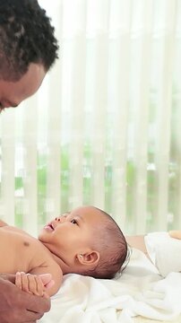 An adult gently holds a baby's hands as the infant lies on a bed, sharing eye contact and a playful, loving moment in soft daylight near a window.