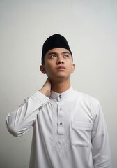 Obraz premium Young Muslim Man in Traditional White Baju Melayu and Songkok Looking Up Thoughtfully Against White Background