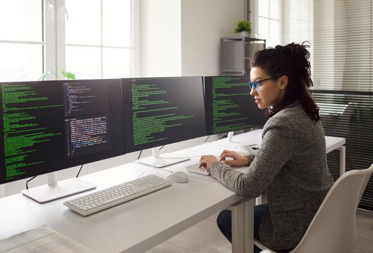 Programmer woman coding software on multiple monitors. Skilled developer in office writes code at computer, debugging bugs, boosts flow with technology. Clear focus on productivity concept.