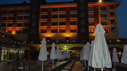 Naklejka premium Panoramic evening view of a luxury resort hotel with illuminated architecture, swimming pool and closed umbrellas under a dark blue sky