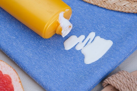 Close-up of white sunscreen lotion spilled from a yellow bottle onto blue fabric with summer accessories nearby. Concept cosmetic stains on the cloth. top view