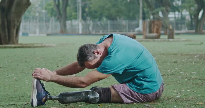 Man with a lower limb prosthesis performing a seated hamstring stretch on grass in a public park, emphasizing adaptive exercise, physical therapy, mobility, flexibility, resilience, healthy 