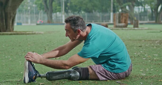 Man with a lower limb prosthesis stretching on the grass before exercise in a public park, promoting adaptive fitness, functional mobility, flexibility, independence, resilience, injury 