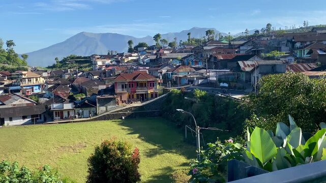 Scenic morning view of a densely populated rural village situated on a mountain slope with a green field in the foreground and a majestic mountain peak under a clear blue sky in Indonesia.