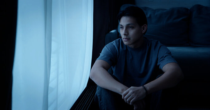 Alone man sit floor by window at night. Sad young person looking out in dark room at home. Gloomy atmosphere, loneliness, depression, solitude. Melancholy mood, introspection, psychological struggle.