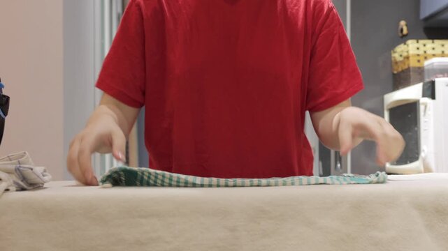 Hands straighten a checked kitchen towel on an ironing board and then guide a steam iron across the fabric.
