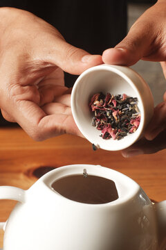 Close-up of hands pouring a blend of loose leaf herbal tea with rose petals from a small ceramic cup into a white teapot filled with hot water.