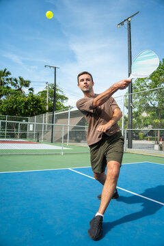 Action shot of male player hitting yellow ball with paddle in mid air on outdoor pickleball court, showing full body active sport motion match gameplay recreation