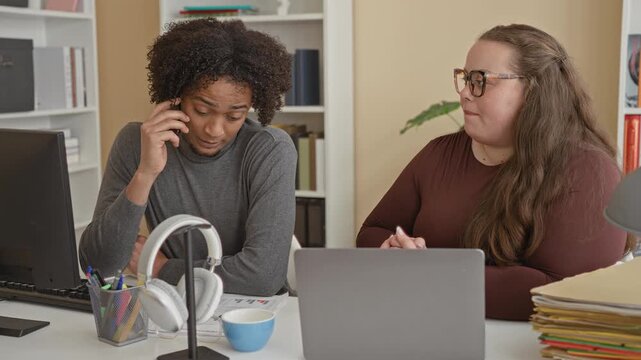 Man on phone and woman gesturing by laptop at a cluttered desk with headphones and folders in a building; frustration teamwork.