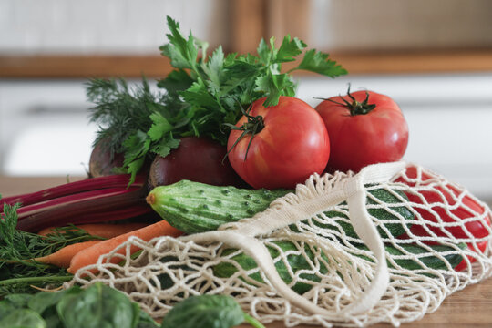 Fresh organic farm vegetables in reusable mesh shopping bag on wooden kitchen table. Sustainable lifestyle and zero waste concept featuring locally grown produce