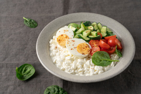 Fresh high protein savory cottage cheese bowl with cherry tomatoes, cucumber, boiled egg and chili flakes served on dark gray linen fabric background with copy space