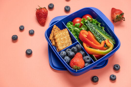 Blue school lunch box with berries crackers and fresh vegetables on coral background, top view back to school healthy snack concept
