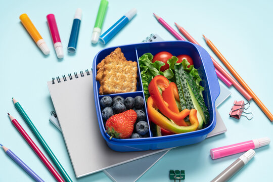 Blue school lunch box with berries crackers and vegetables surrounded by stationery on blue background, top view back to school concept