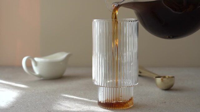 Black coffee pouring from glass carafe into clear glass cup on light background, slow motion morning drink preparation concept