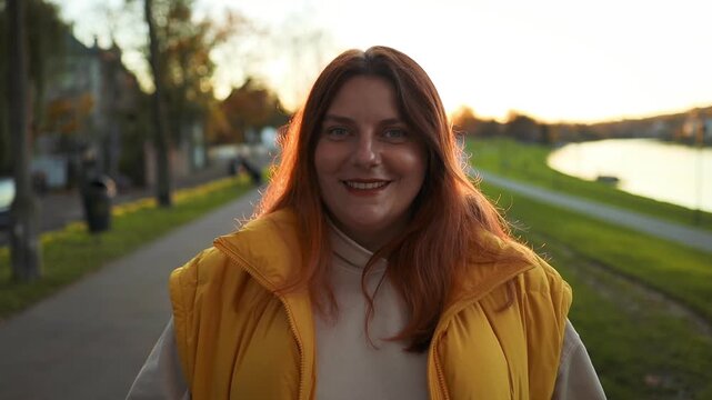 A woman is smiling and standing still on an outdoor pathway with a river in the background. She appears happy and relaxed, captured from a frontal view slightly elevated. Her yellow and/or.