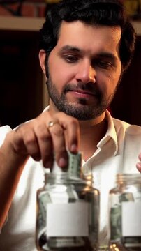 Indian bearded young man sitting at a desk under moody lighting, managing finances by putting money into two glass jars with blank labels for customisation, symbolising saving and financial planning