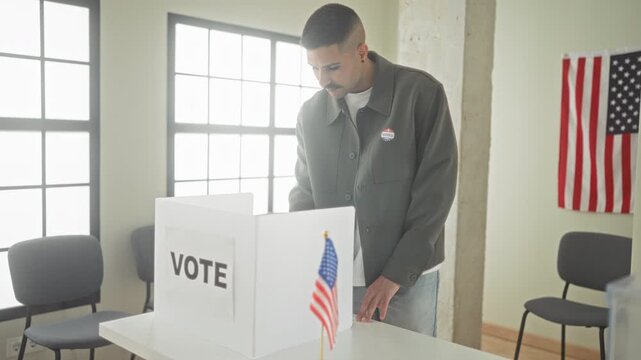 Man places ballot into voting booth at polling building while wearing a voted sticker and standing beside an american flag; civic pride.