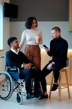 Vertical shot of diverse coworkers discussing smartphone content during office meeting, Black man with disability using wheelchair. Supporting inclusion, accessibility, workplace diversity campaigns