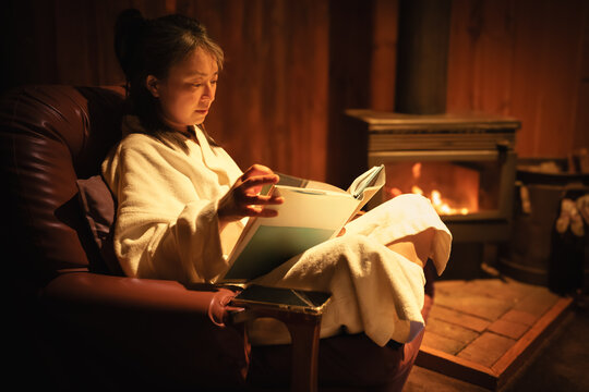 Side view of a woman wrapped in a white robe sits in an armchair, reading a book while a wood burning fireplace glows in the background, Mount Macedon, Victoria, Australia