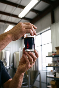 a brewer s hands holding a small tasting glass of dark stout, inspecting the color against bright warehouse lighting