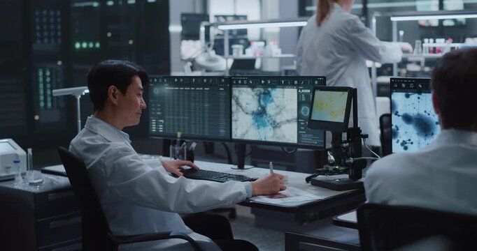 Asian Male Researcher Reviews Microscopy Scans and Molecular Data on Multi-screen Workstations as Colleagues Test Samples in a Modern Medical Biotech Lab Advancing Drug Discovery and Disease Treatment
