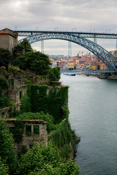 Dom Luis I Bridge spanning the Douro River in Porto Portugal with ivy covered ruins on the riverbank and historic city buildings beyond