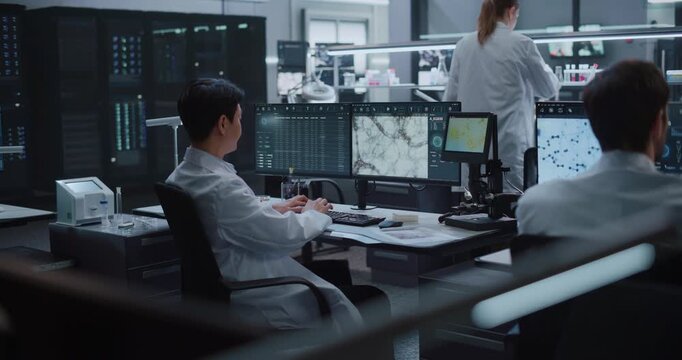 Asian Male Scientist Logs Microscopy Images and Cellular Datasets on Monitors While Lab Team Processes Samples in Biotech Center for Drug Development, Diagnosis and Disease Therapy Research