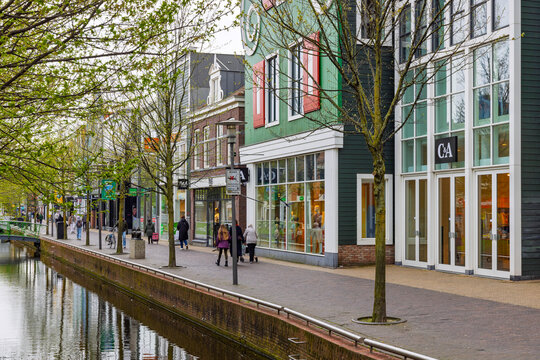 Zaandam, Netherlands - 11 April 2026 - Pedestrians walking along a scenic canal lined with modern shops featuring traditional green Zaans architecture on a cloudy day.