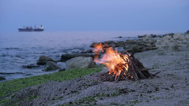 Bonfire burning on a rocky beach at dusk with cargo ship on the horizon