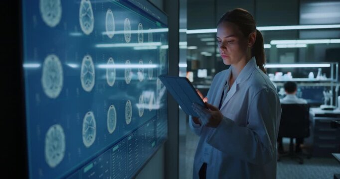 Young Caucasian Woman Researcher Uses Digital Tablet to Study MRI Brain Scans Displayed on a Large Interactive Monitor Inside Biotech Laboratory, Advancing Neuroscience and Treatment Innovation
