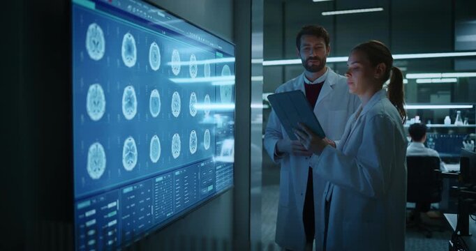 Male and Female Neurologist Medical Scientists Talk in Meeting, Examining MRI Brain Scans on a Large Touchscreen Display While Using a Tablet in Medical Research Hospital Laboratory, Drug Development