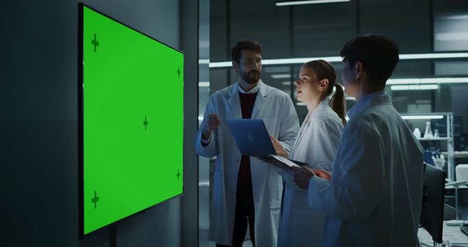 Diverse Team of Medical Scientists Meeting and Talking Near a Large Green Screen Chroma Key Display, Holding Laptop and Tablet in Hospital Research Laboratory Advancing Neurology Drug Development.