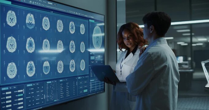 Black Female Neurologist and Asian Male Doctor Meeting and Talking, Examining MRI Brain Scans on a Large Interactive Display With Laptop in a Modern Hospital Lab, Researching Neuroscience Drug Therapy